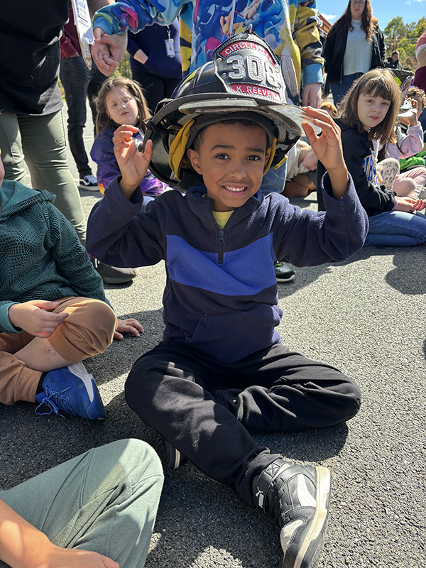 An elementary age boy sits on the ground. He is wearing a black shirt with blue stripe across it and he is smiling. He has a big fire helmet on his head.