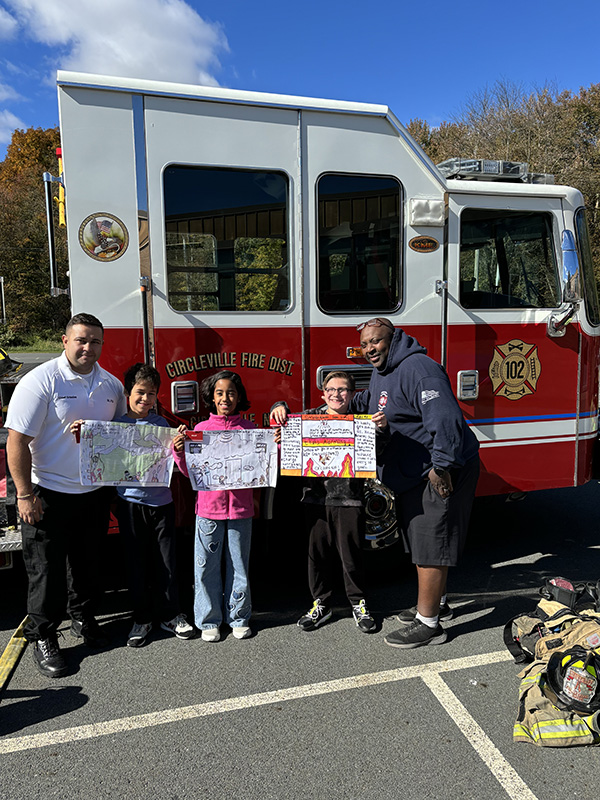 Three elementary students stand holding their posters. They are in front of a red and white fire truck. Next to them are a fire fighter on either side. 