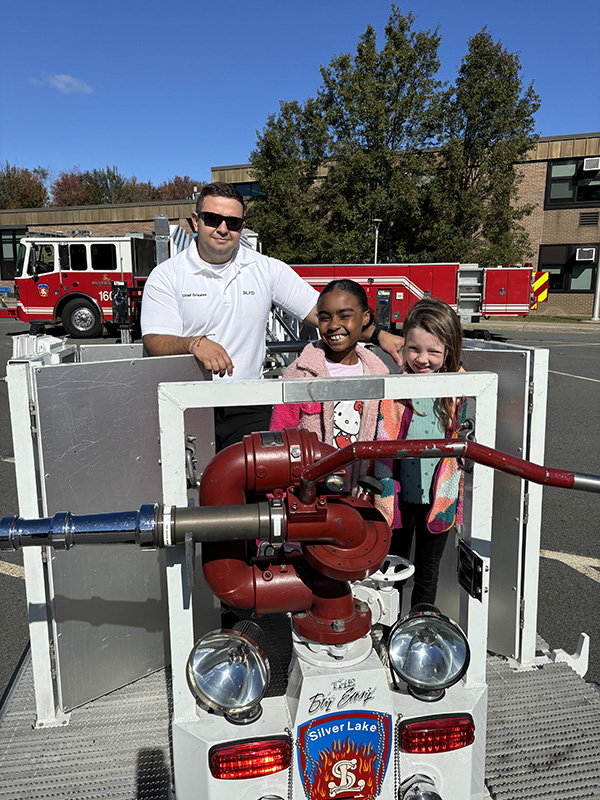 Two elementary girls stand on top of a fire truck with a firefighter standing with them.