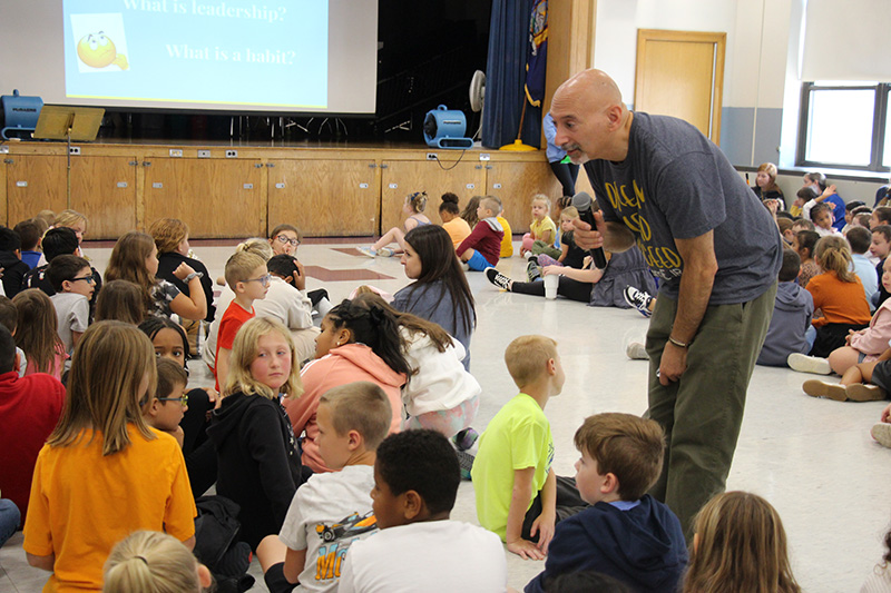 A man holding a microphone bends down to talk to smaller elementary students.