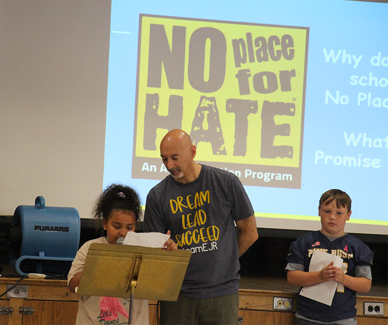 A man in a blue t shirt that says Dream Lead Succeed listens as an elementary student reads her essay for a group of kids.