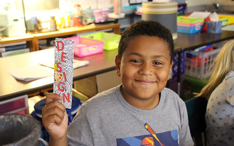 A fourth grade boy with short dark hair smiles holding up the bookmark he colored that says DESIGN.