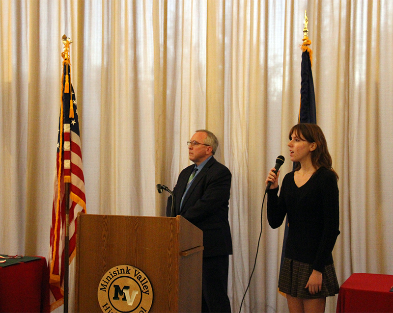 A young woman on the right is holding a microphone and singing the National anthem. There is a man to the left and an American flag on the far left.