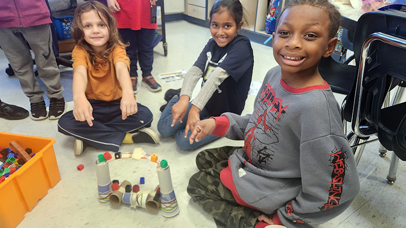 A group of three second-grade students smile as they sit on the floor working on building a bridge.