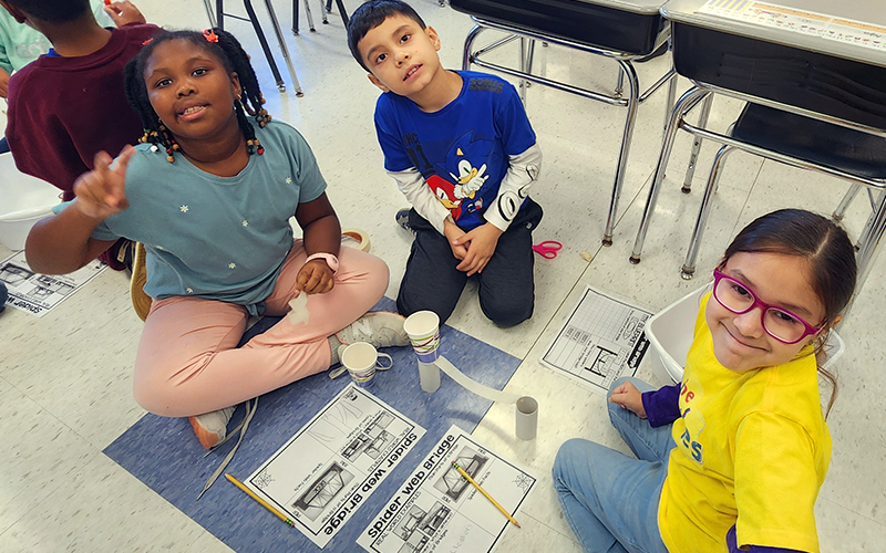 Three second-grade students sit on the floor and smile as they build a bridge.