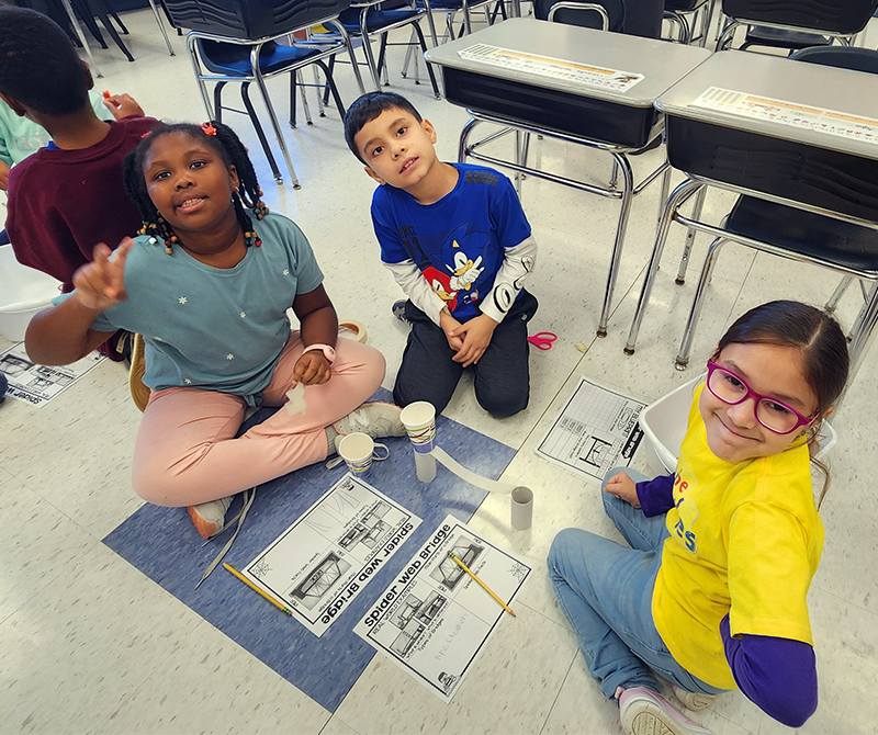 Three second-grade students sit on the floor and smile as they build a bridge.