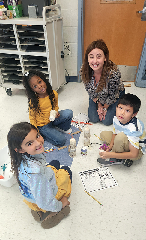 Three second-grade students sit on the floor smiling. They are with a teacher as they build a bridge from different materials.