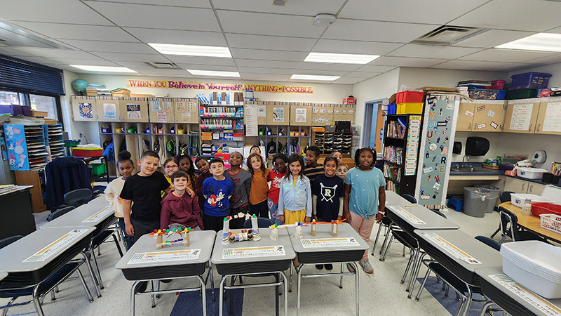 A class of second-graders stand behind tables that have the bridges they built.