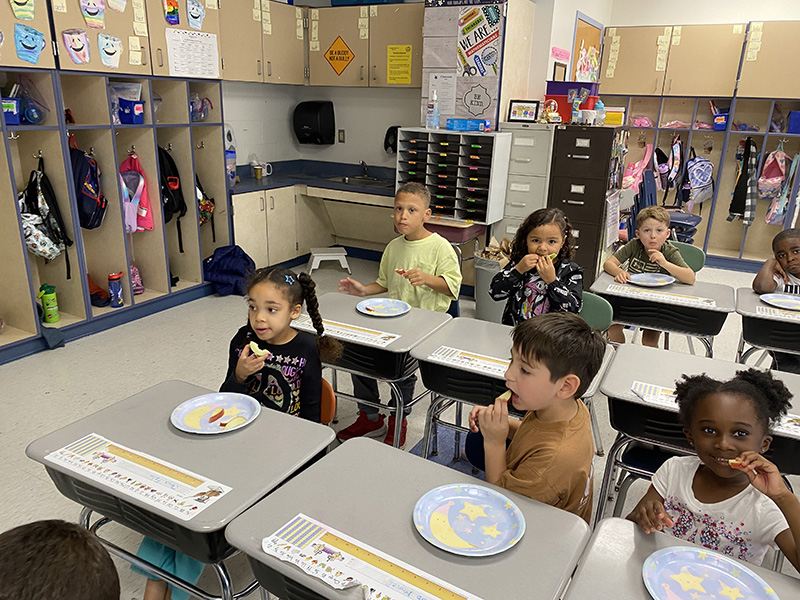 Six first grade students sit at their desks with paper plates in front of them. On the plates are slices of apple. They are tasting them.