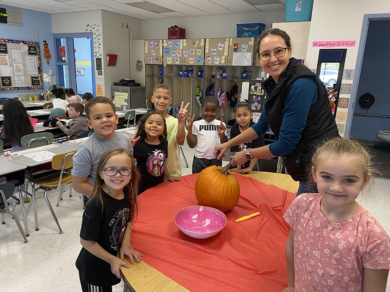 A woman with dark hair smiles as she cuts open a pumpkin. Around her are five first-grade students smiling and waiting.