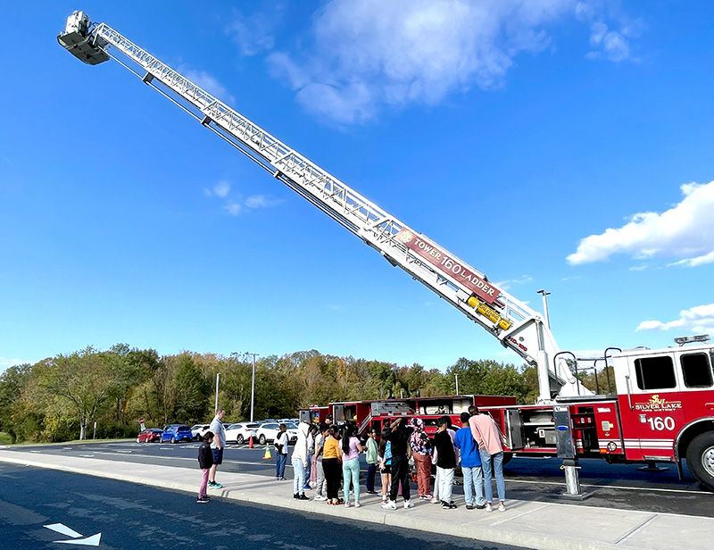 A bright blue sky in the background. A class looks up at a very tall ladder extended on a fire truck.