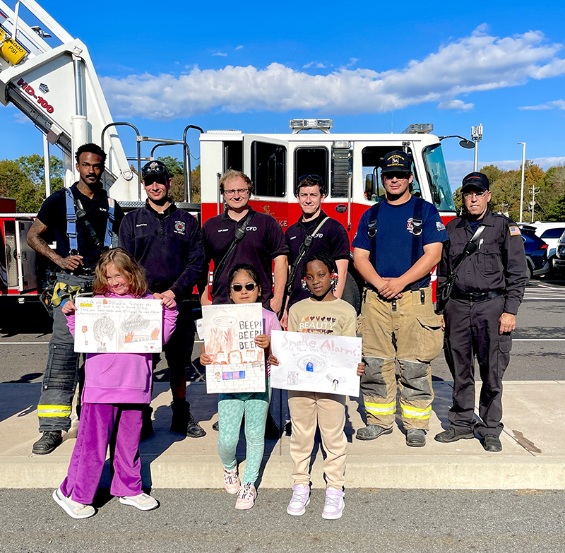 Three third grade students hold up their posters. Behind them are six fire fighters. and a large red fire engine.