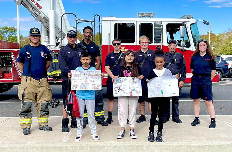 Three fourth-grade students hold up the posters they created for fire safety week. Behind them are six fire fighters and a fire truck.