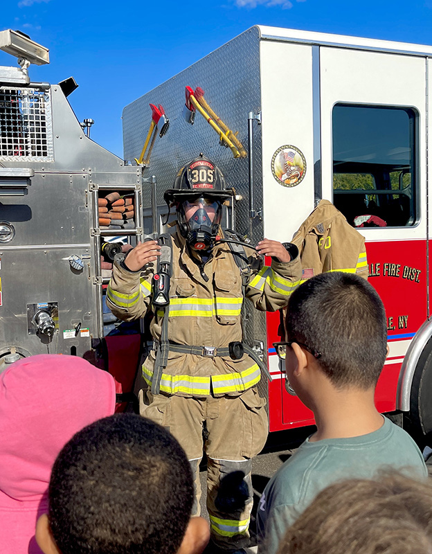 A fire fighter dressed in his fire equipment talks to a group of fourth-grade students.