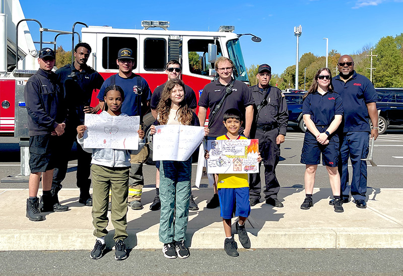 Three fifth-grade students stand with the posters they created for fire safety week. Behind them are six fire fighters and a fire engine.