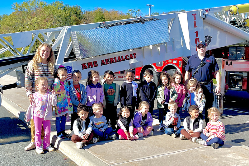 A class of second grade students and their teachers stand in front of a large fire truck with some fire fighters.