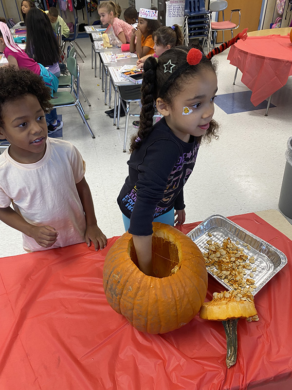 Two first-graders stand near a table with a pumpkin on it. One girl has her hand in the top of the pumpkin.