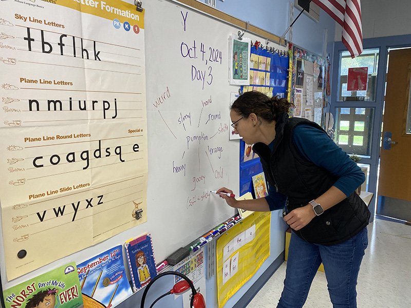 A woman with dark hair pulled back writes on a large sheet of white paper hanging on a board.