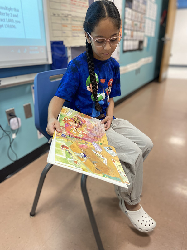 A girl wearing a bright blue shirt and glasses sits on a chair with a book turned toward a class of kids.