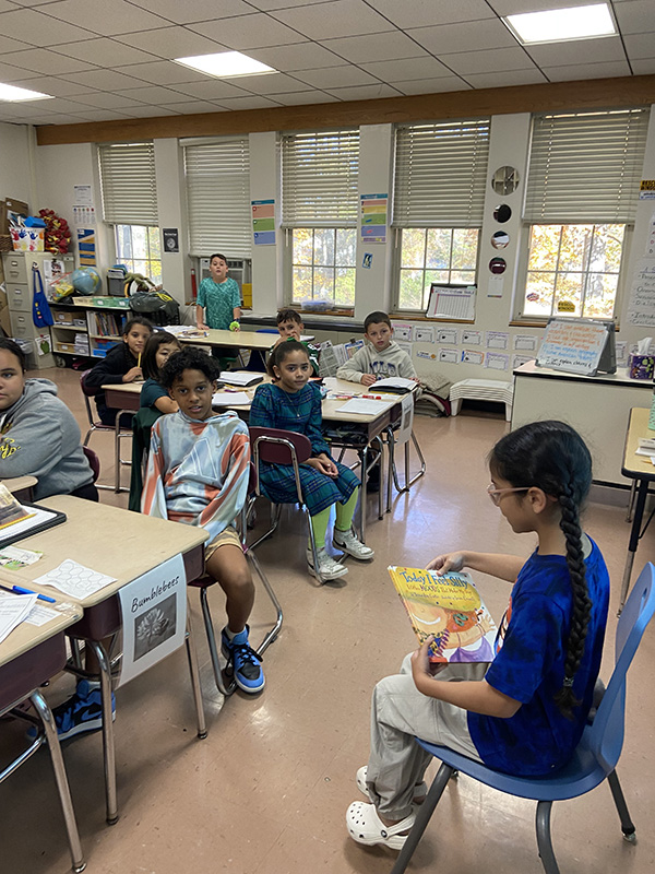 A girl with long dark hair, wearing a bright blue shirt, turns the page on a book she is reading to a classroom filled with kids.