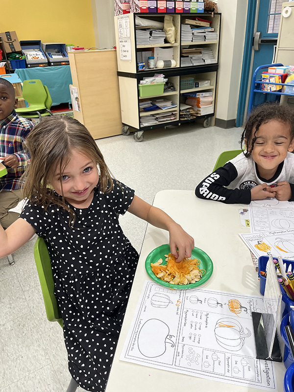 A little girl puts her hand in a plate filled with pumpkin seeds and flesh. Another kid watches and smiles.