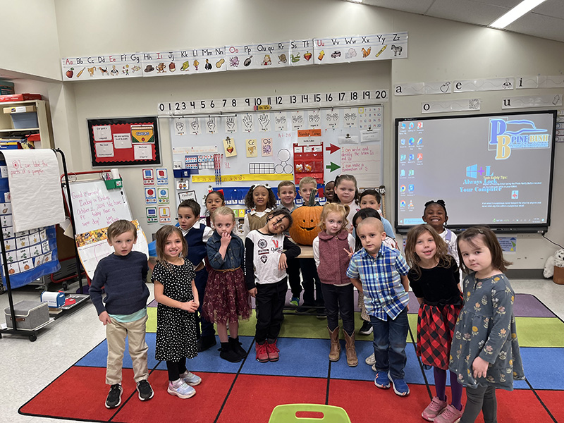 A class of kindergarten kids stand and smile with a carved pumpkin behind them.