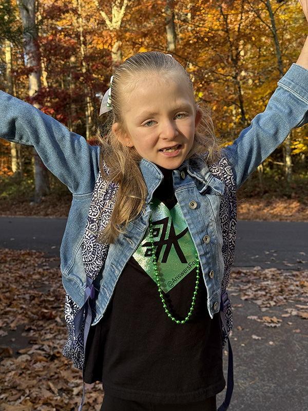 A little girl withlong blonde hair holds her hands up in the air and smiles. She is wearing a denim jacket and a black and green shirt.