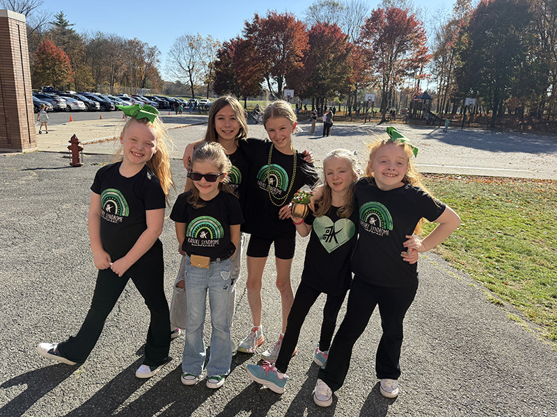 A group of six young elementary age girls stand outside on a beautiful fall day. They are all wearing black t shirts with green arches on them.