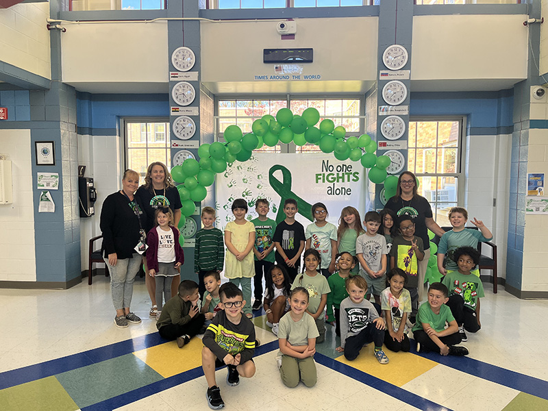 A group of 22 elementary students and three adults stand under an arch of green balloons.