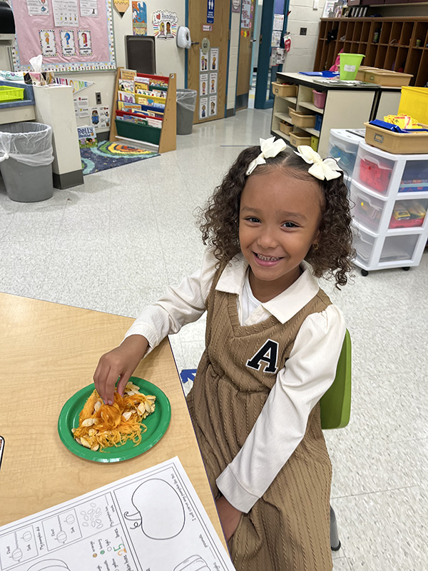 A little girl with white bows in her hair, wearing a tan shirt and white blouse smiles as she touches the inside of pumpkin on a plate.