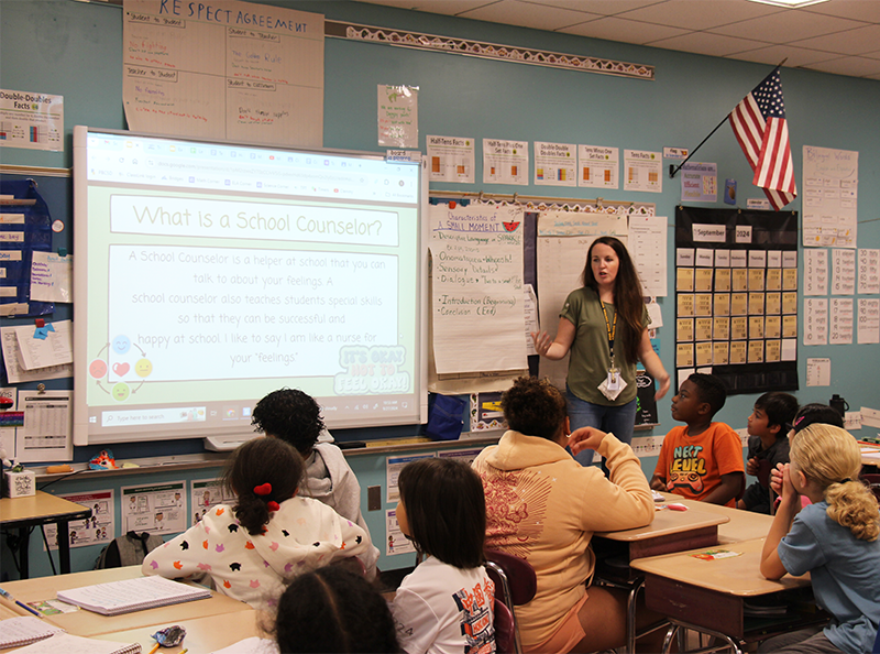 A woman with long dark hair stands in front of a class of fourth-graders, teaching.