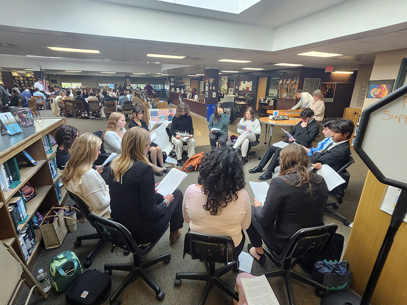 A group of high school students sit  in a circle and listen as other students present.