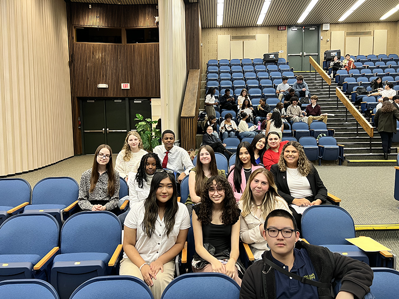 A group of 12 high school students sit in an auditorium smiling.