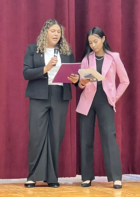 Two high school age young women stand on a stage . Both are wearing professional suits.