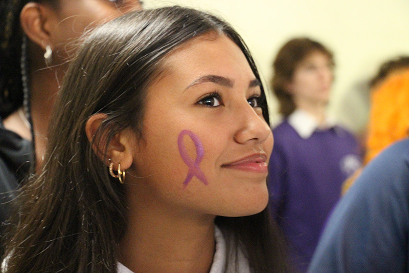 A young woman with long dark hair smiles. she has a purple ribbon painted on her cheek.