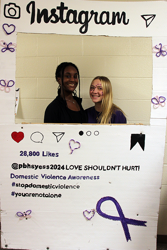 Two high school girls stand inside of a white photo booth. they are smiling and the message is domestic violence prevention and awareness.