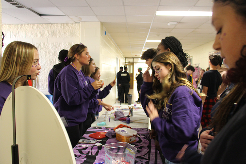 A group of high school students stand on two sides of a table. They are talking and dressed in purple.