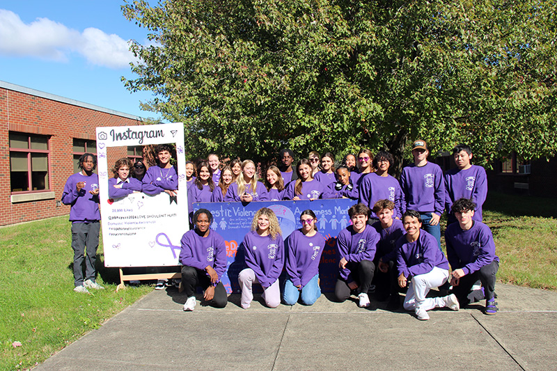 A large group of high school kids - about 30 - all wearing purple sweatshirts, stand together smiling. There is a tree and blue sky in the background.