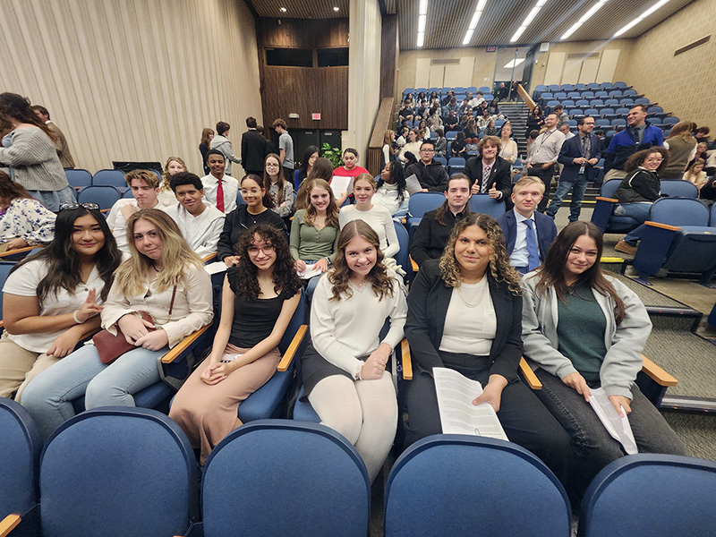 A group of about 25 high school students sit in an auditorium smiling.