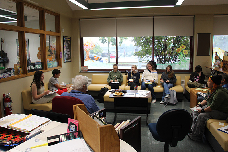 Large windows in the background, there are a group of about 8 high school students witting on couches and chairs with three adults. On the table in the center, are cookies.