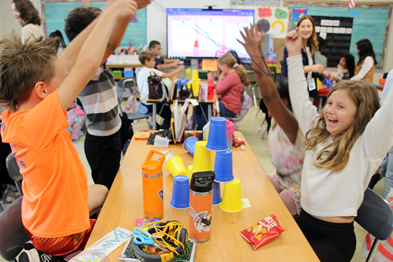 Happy fourth-graders throw up their arms and smile after finishing the pyramid of cups.