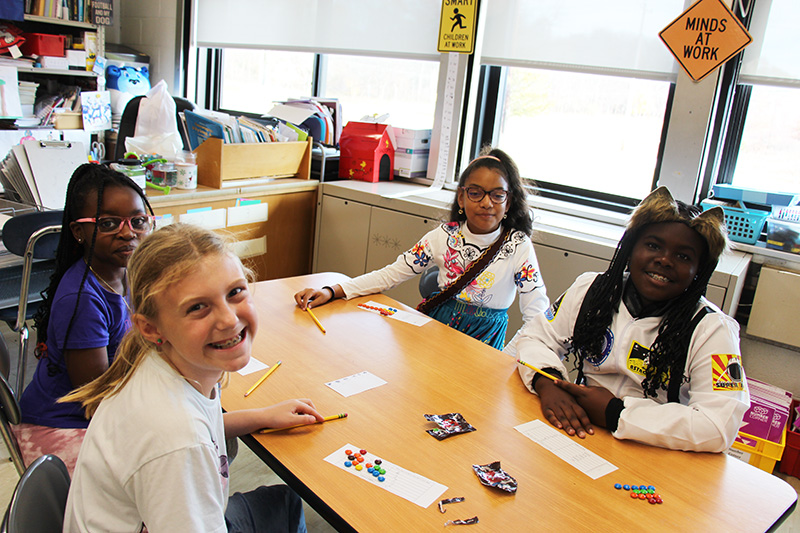 A group of fourth-grade girls sit around a table and smile. There are papers and multi-colored M&Ms on the table.