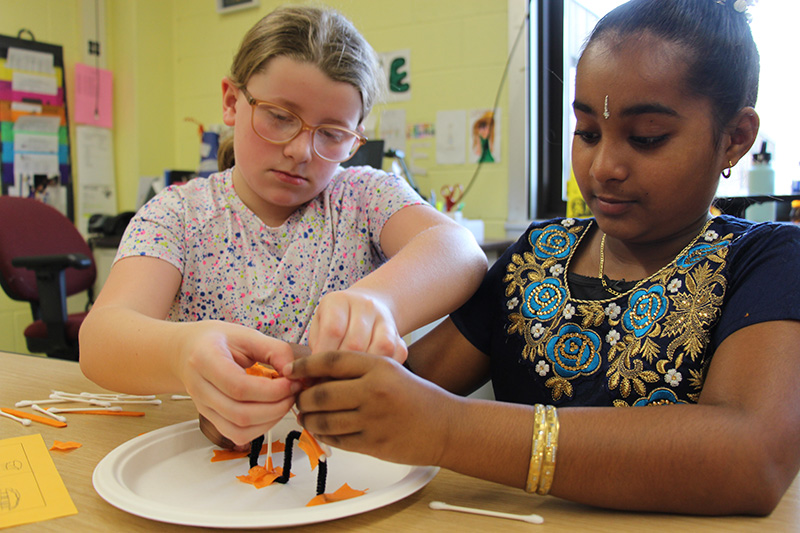 Two fourth-grade girls work on creating a bridge out of Q-tips, tape and pipe cleaners.