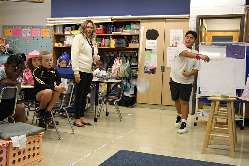 A fourth-grade boy in a white tshirt and dark shorts lets go of a paper airplane as a class full of peers watche.