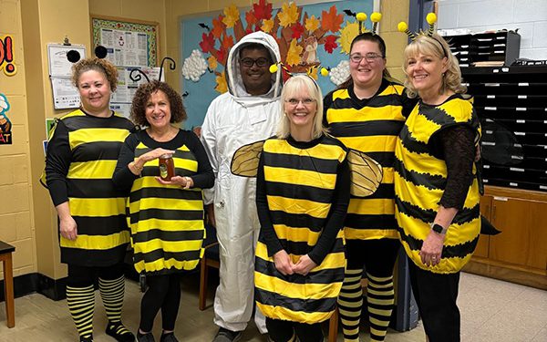 Six adults are smiling in an office. Five women are dressed as yellow and black bees. The man is dressed in a white coverall as a beekeeper.