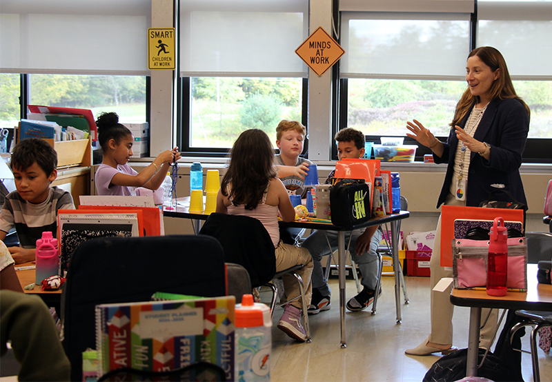 A classroom of fourth-grade students, some with hands up, as a woman speaks to them.