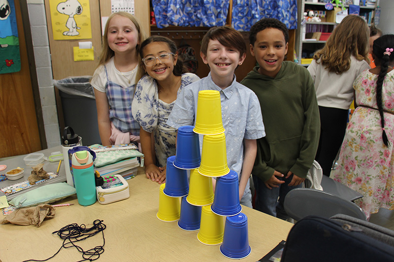 Four fourth-grade students smile as they stand behind a table. In front of them are blue and yellow cups stacked in a pyramid.