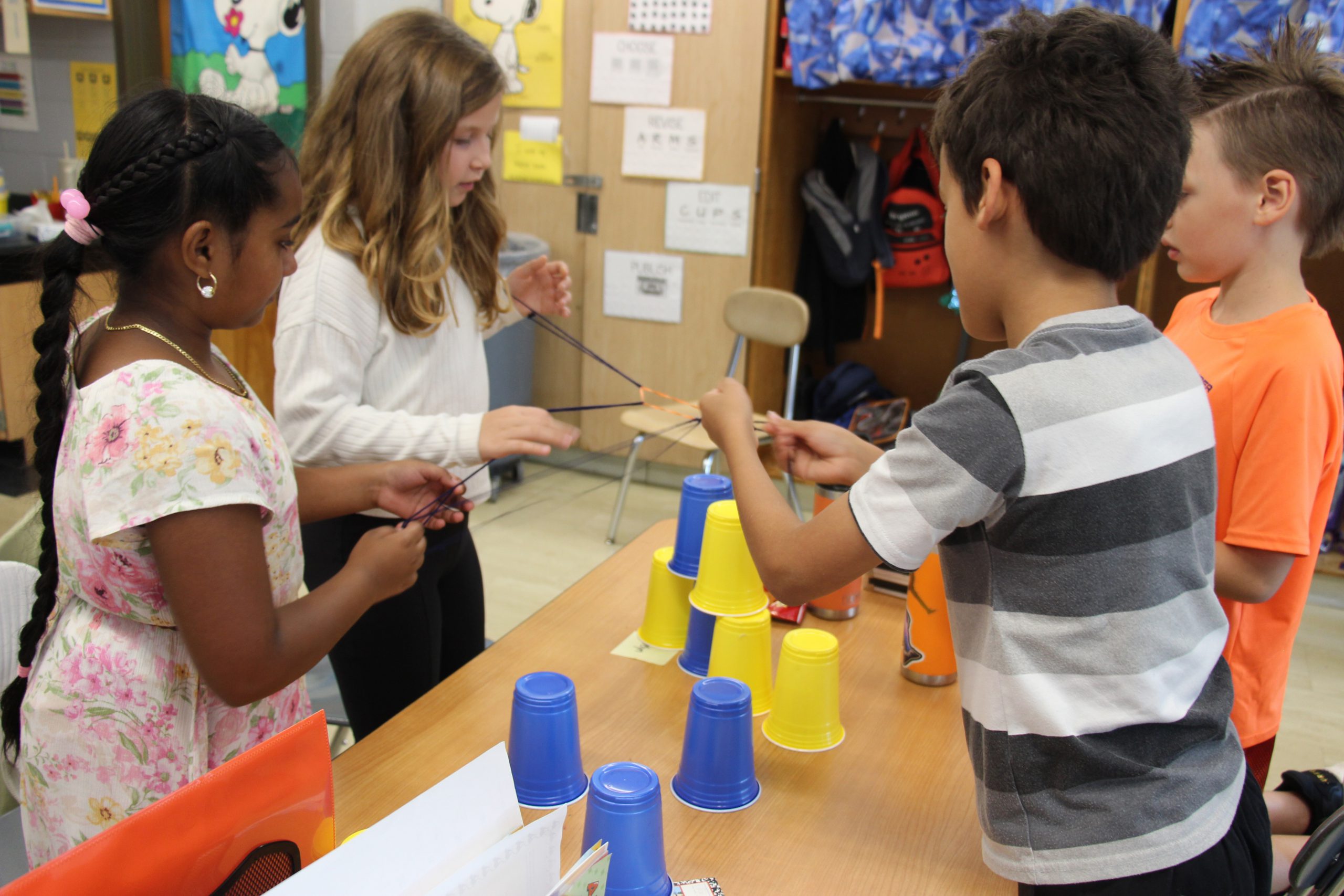 Four fourth-grade students uses an elastic with handles to stack blue ad yellow cups in a pyramid.