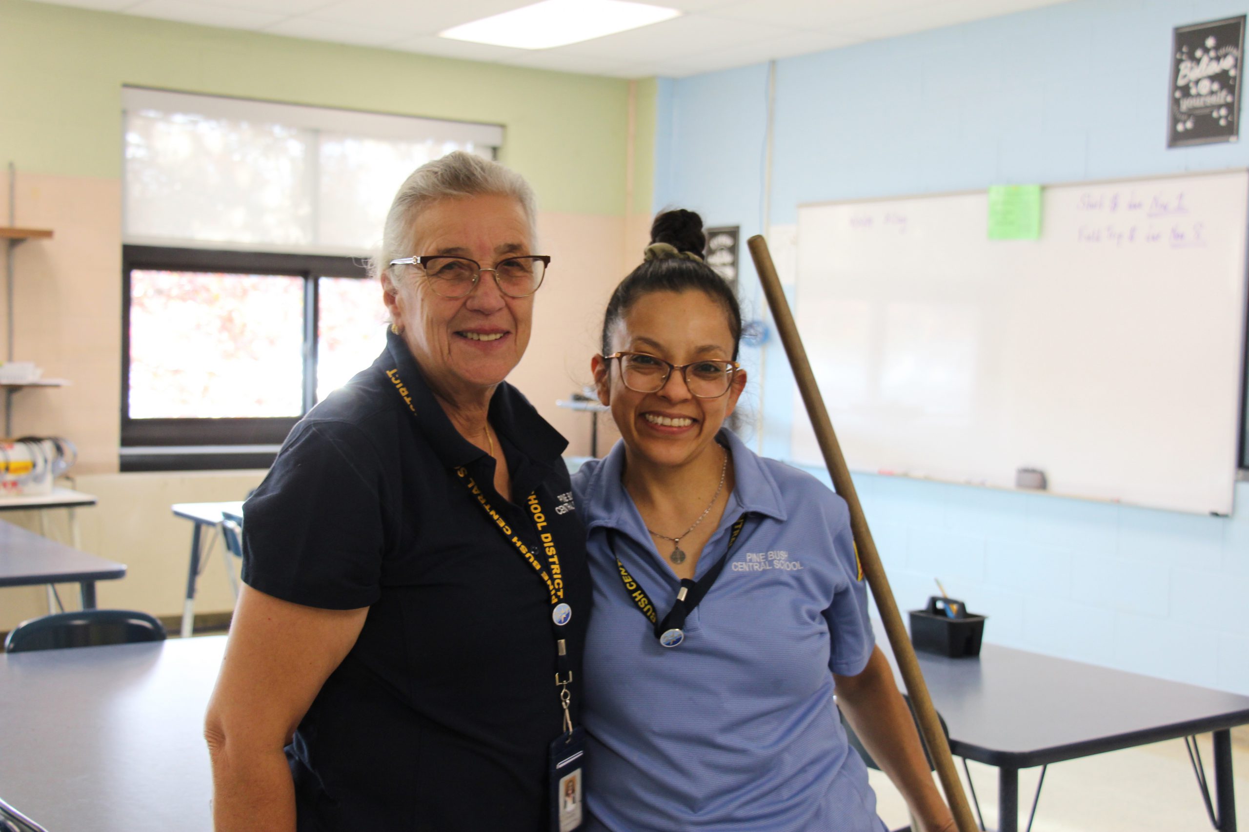 Two women, both with their hair pulled back, smile. They are wearing blue shirts and one is holding a broomstick.