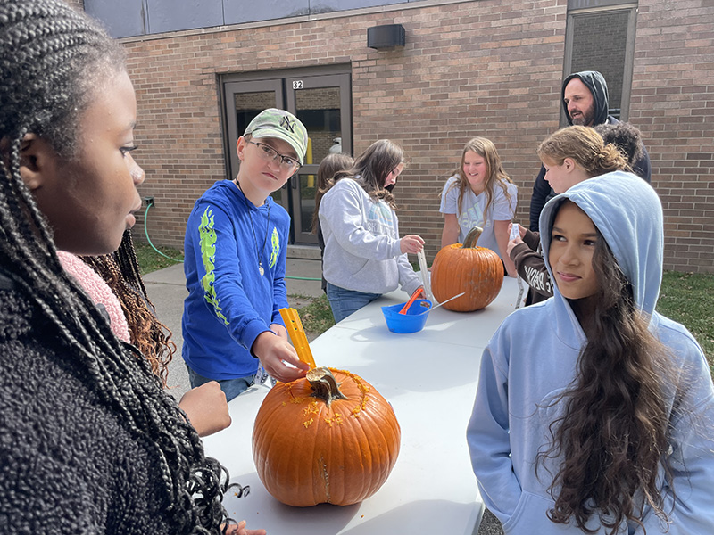 Middle school kids at a table with pumpkins. They are measuring them and taking the top off.